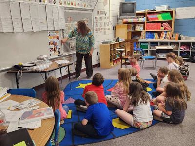 Elementary teacher standing at whiteboard pointing to poster while students sit on rug