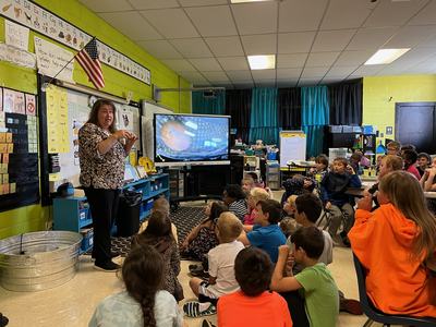 Teacher gesturing while students sit on floor watching a screen in a classroom
