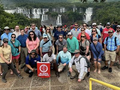 A group poses with an NC State University banner in front of a large waterfall.