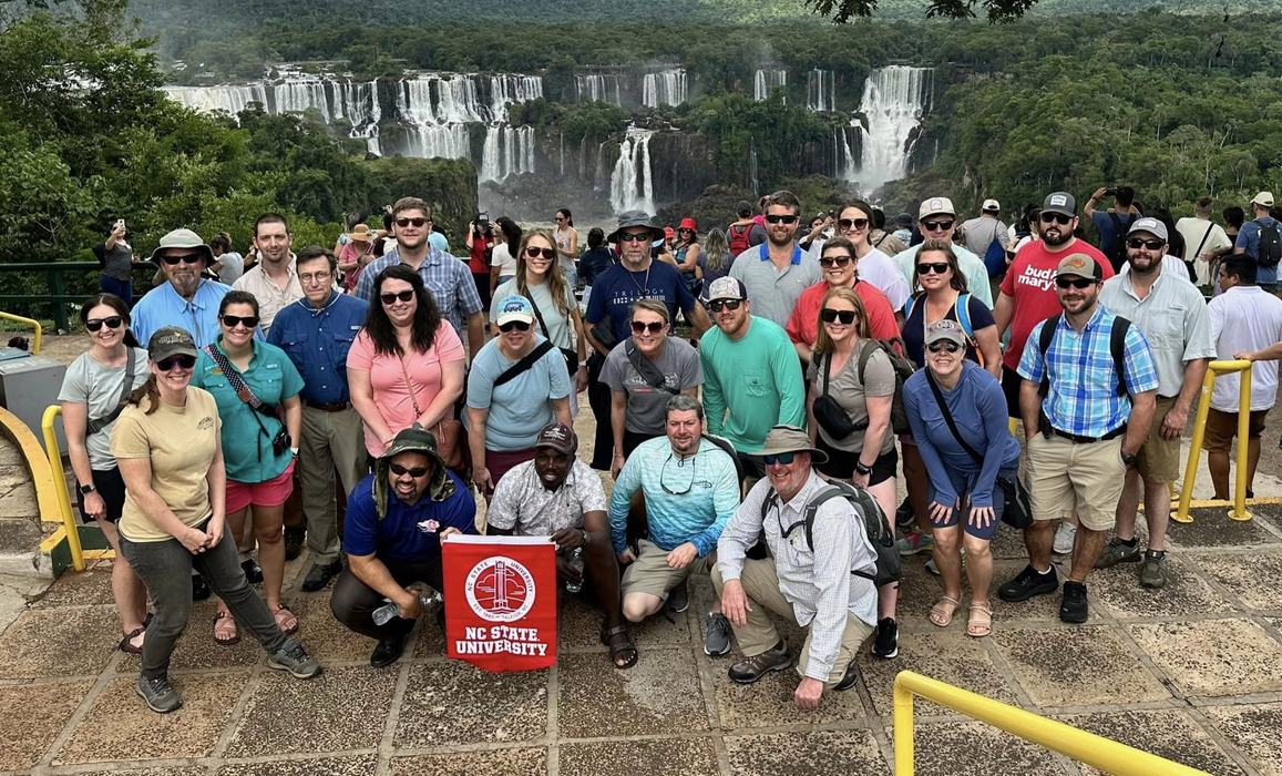 A group poses with an NC State University banner in front of a large waterfall.