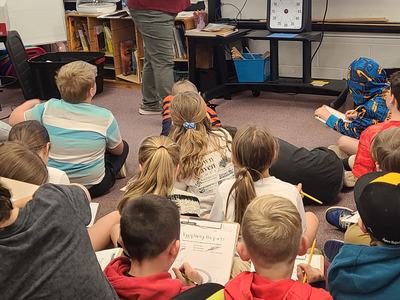 Teacher points to egg diagram on screen while students sit on floor writing.