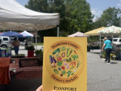 Hand holding "Henderson County Farmer's Markets PASSPORT" at outdoor farmers market