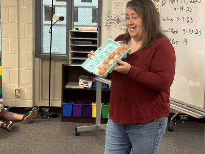 Woman holding an open egg carton with eggs in front of a classroom whiteboard dated April 19, 2023