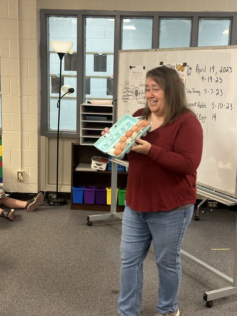 Woman holding an open egg carton with eggs in front of a classroom whiteboard dated April 19, 2023