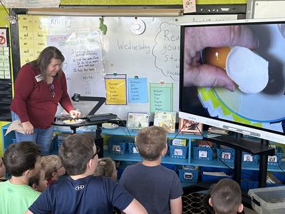 Teacher demonstrating cracking an egg on a document camera while students watch a large screen