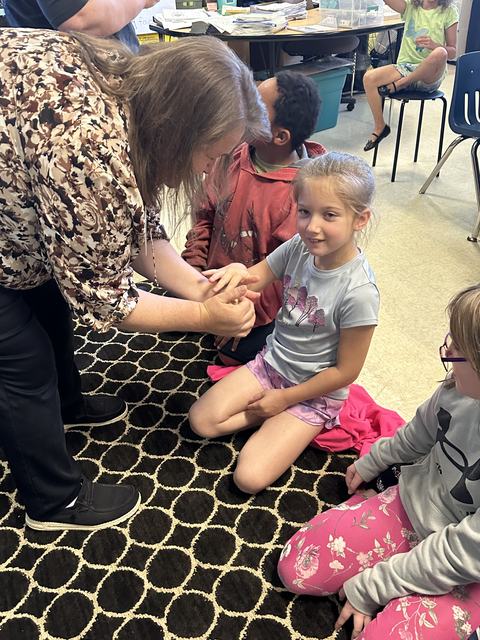 Adult bending over to hold a seated girl's hands while other children sit nearby