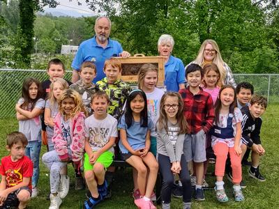 Group of elementary students with teachers displaying a wooden observation hive with bees outdoors