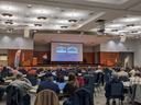 Presenter on stage giving a slideshow to a seated audience in a conference hall