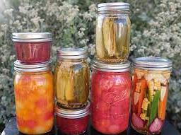 canned-food-in-jars.jpeg seven mason jars with tomatoes in one, pickle spears in two, red jam in two, a variety of vegetables in one jar with corn and other vegetables, and fruits in one jar.