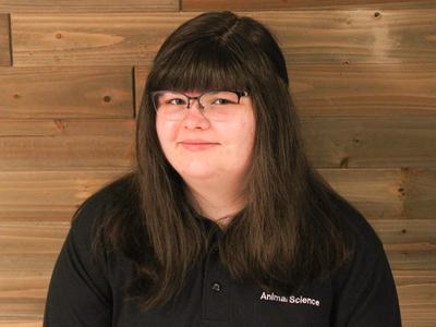 Person with long dark hair and glasses wearing black polo labeled "Animal Science" against wood