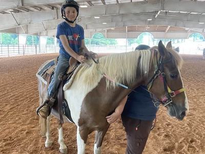 Child in helmet sitting on brown-and-white horse in indoor riding arena, handler beside horse