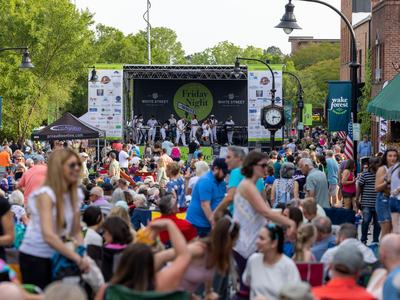 A crowd of people are sitting and standing in a downtown street that leads to a stage with a musical group performing in front of Friday Night on White sign.