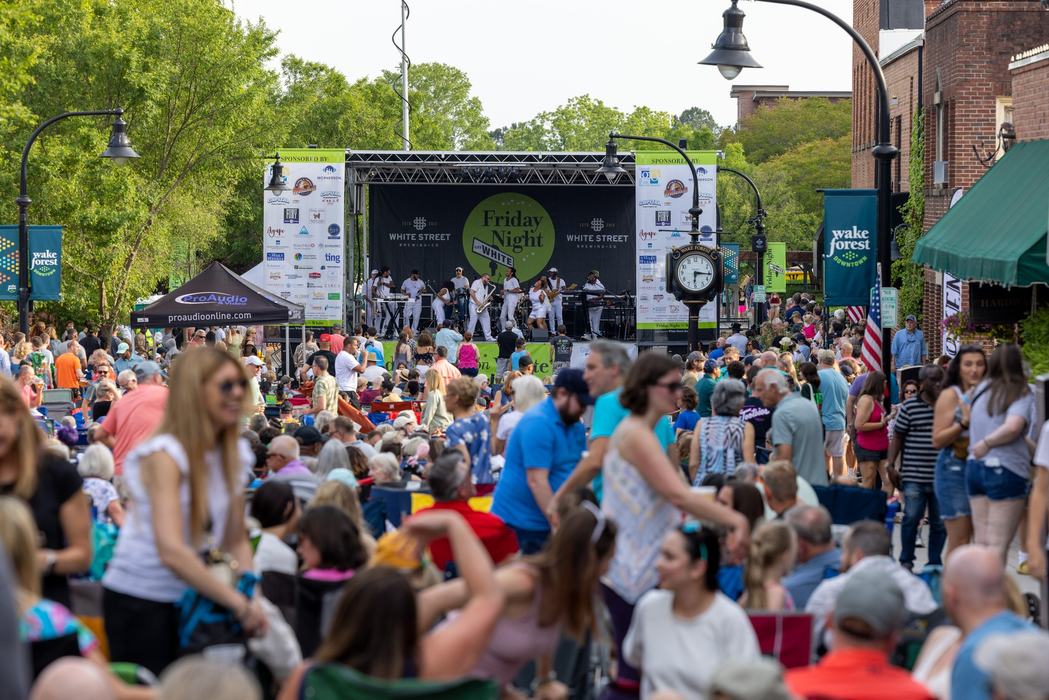 A crowd of people are sitting and standing in a downtown street that leads to a stage with a musical group performing in front of Friday Night on White sign. 
