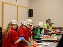 Children in chef hats and aprons preparing food at a long table in a cooking class