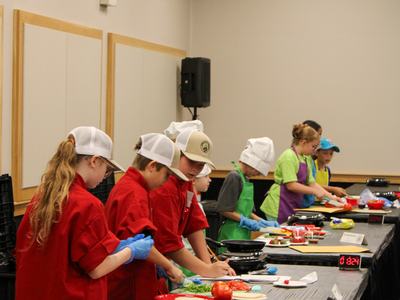 Children in chef hats and aprons preparing food at a long table in a cooking class
