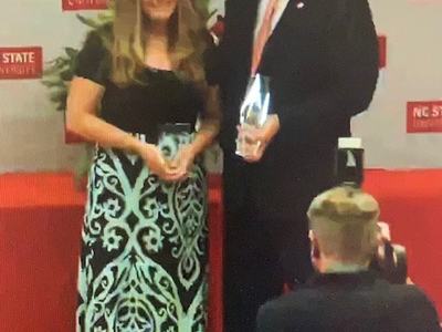 Two people holding awards on stage with backdrop reading "NC STATE UNIVERSITY."