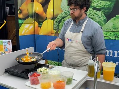 Cook stirring diced vegetables in a frying pan at a demo counter with prep bowls