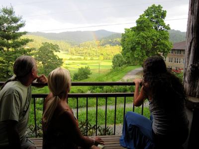 Three people on a porch railing looking at a rainbow over a green valley
