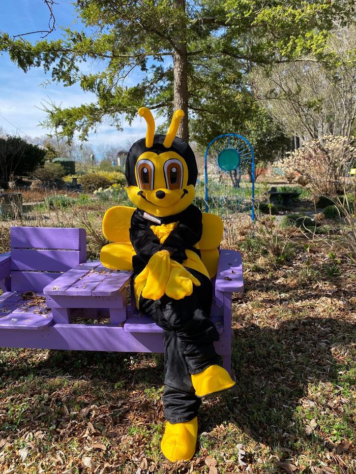 Person in yellow-and-black bee mascot costume sitting cross-legged on purple bench in garden