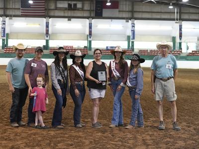 Group of people in western wear posing in indoor arena, center woman holding a plaque