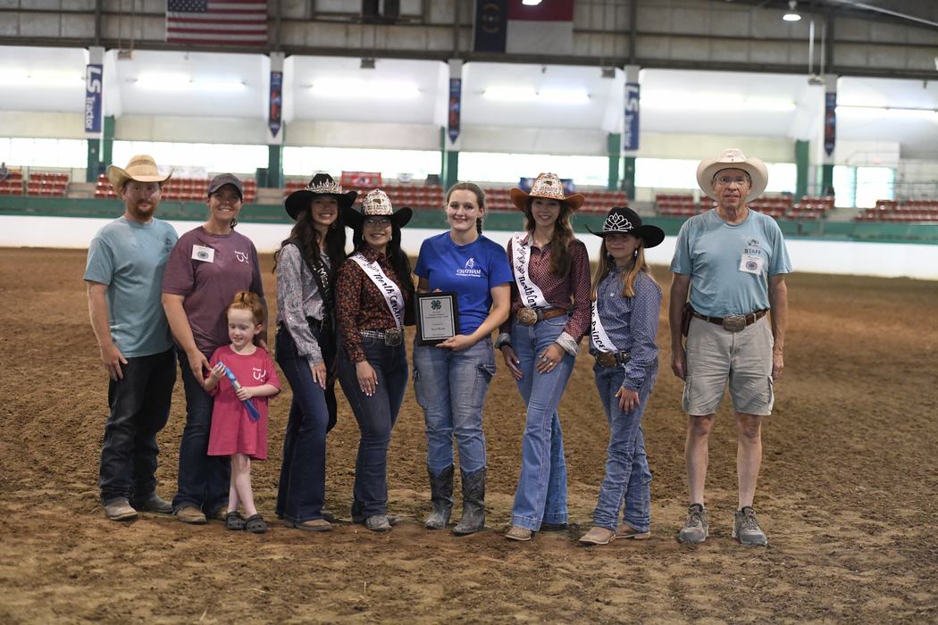 Group of rodeo contestants and staff in arena, woman holding plaque