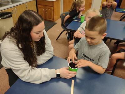 Adult supervising child pressing soil into green cup at classroom table