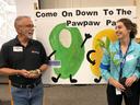 Two people holding a pawpaw fruit and a sapling before sign "Come On Down To The Pawpaw"
