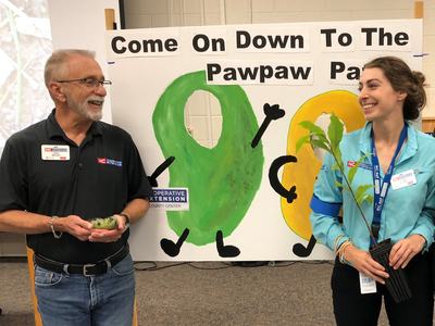 Two people holding a pawpaw fruit and a sapling before sign "Come On Down To The Pawpaw"