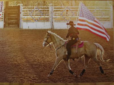 Cowboy-hatted rider carrying American flag while trotting palomino horse in arena