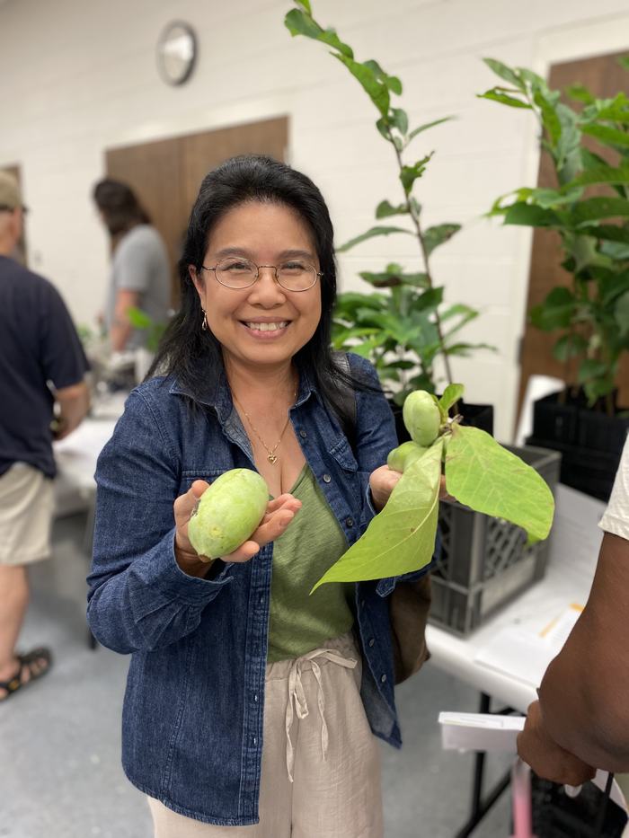 Woman holding two green fruits with attached leaves indoors at a plant event