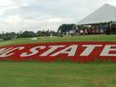 Red turf rectangle reading "NC STATE" on grass beside a tent with spectators