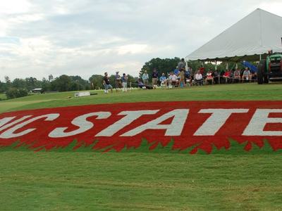 Red turf rectangle reading "NC STATE" on grass beside a tent with spectators