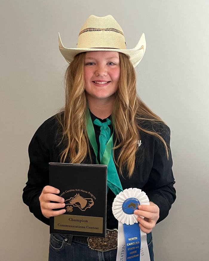 Person in cowboy hat holding "Champion Communications Contest" plaque and NC blue ribbon
