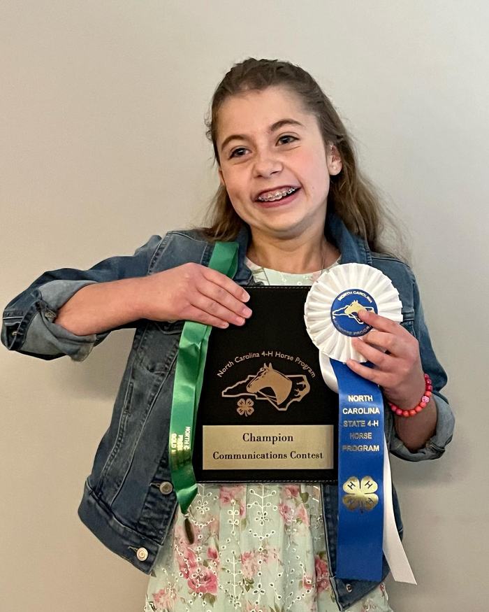 Child holding plaque reading "Champion Communications Contest" and 4‑H blue ribbon