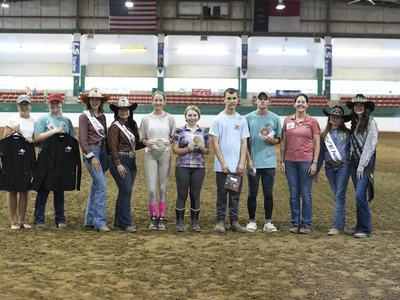 Twelve people lined up in an indoor riding arena holding awards, ribbons, and jackets