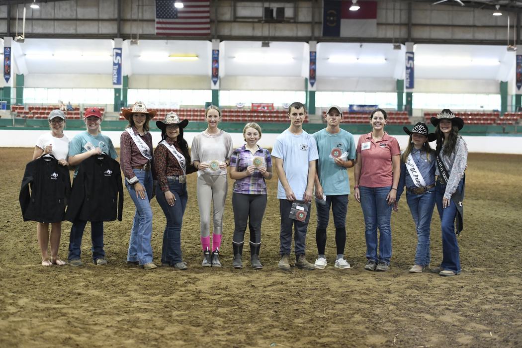 Twelve people lined up in an indoor riding arena holding awards, ribbons, and jackets