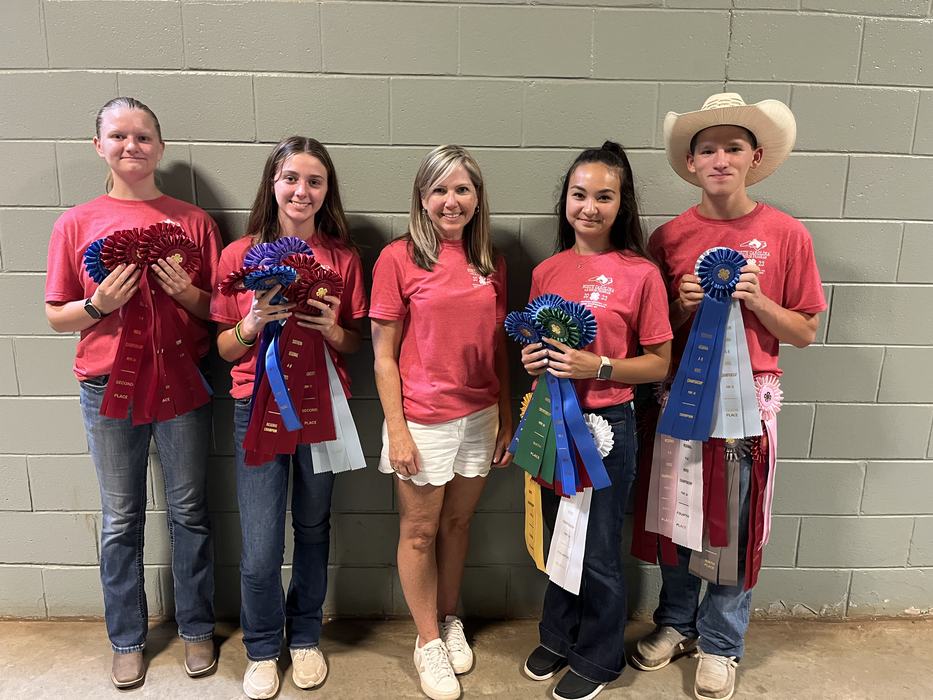Five people in red shirts standing against a cinderblock wall holding many show ribbons.