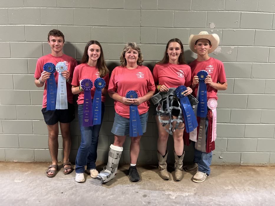 Five people in red shirts holding multiple blue and purple award ribbons against a cinderblock wall