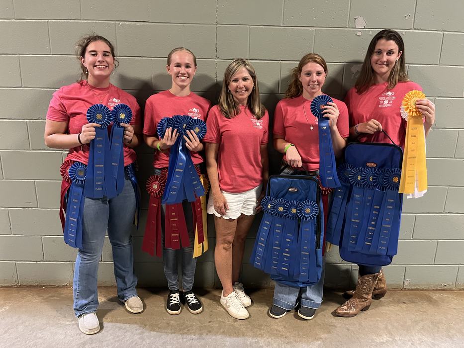 Five people in red shirts holding many blue and yellow award ribbons against a cinderblock wall