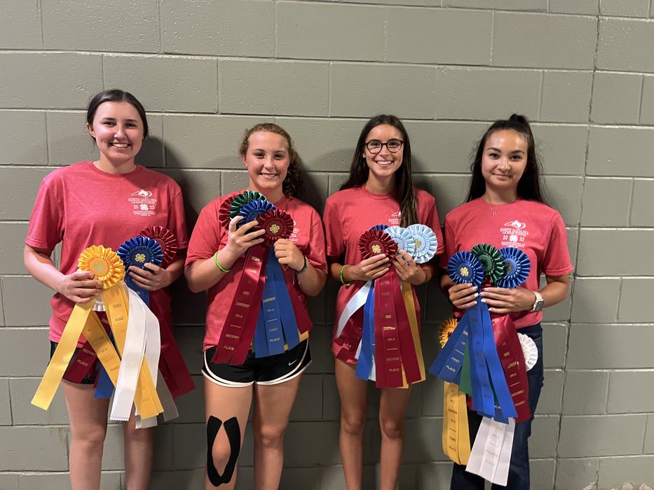 Four girls in red shirts standing against a cinderblock wall holding multiple award ribbons