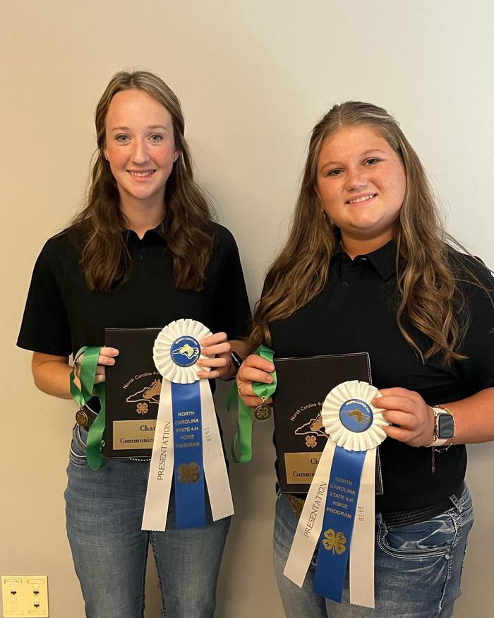 Two young women holding plaques, medals, and blue-and-white ribbons reading "North Carolina State 4‑H Horse Program"