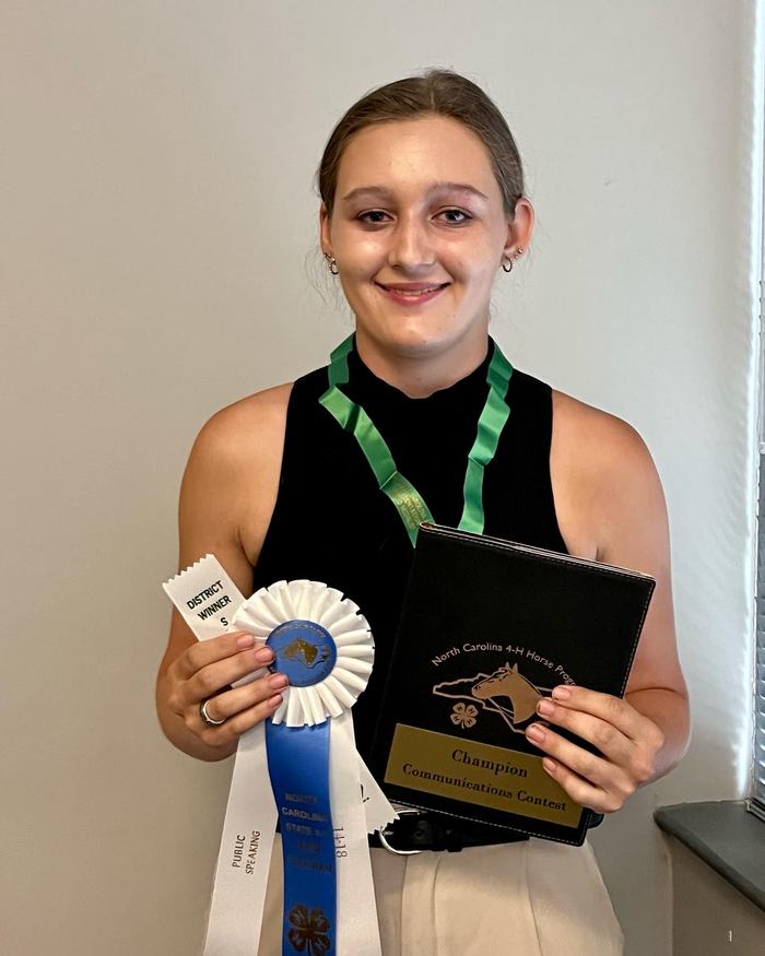 Person holding blue rosette, ribbon reading District Winner, and plaque "Champion Communications Contest"