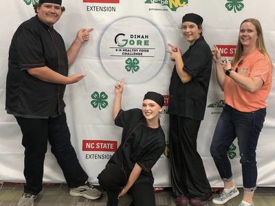 Four people in chef uniforms pointing to sign reading "Dinah Gore 4-H Healthy Food Challenge"