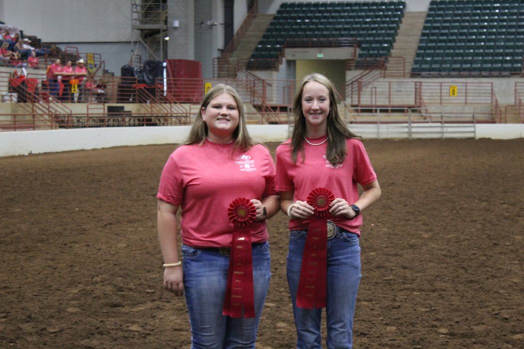 Two people wearing pink shirts holding red rosette ribbons in an indoor dirt arena