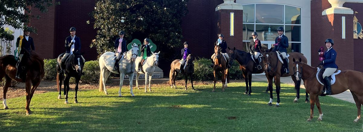 Equestrians on horses holding ribbons outside a brick arena building