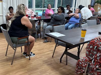 Group of adults seated around tables in a community room, many with arms crossed across their chests.
