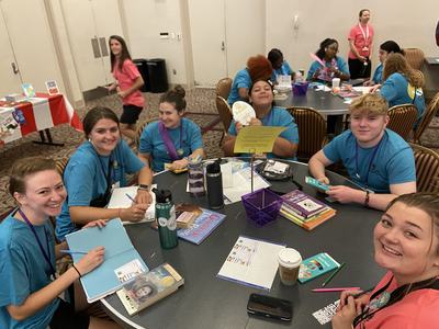 Group of young people in teal shirts seated at round table doing crafts and writing