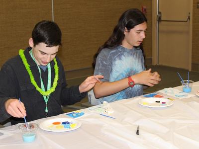 Two adolescents sitting at a table painting on paper plates with brushes and blue paint.