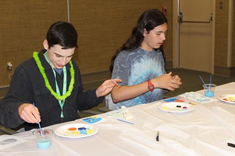 Two adolescents sitting at a table painting on paper plates with brushes and blue paint.