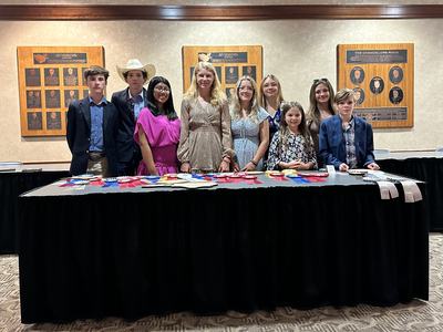 Ten young people standing behind a table covered with award ribbons in a conference room